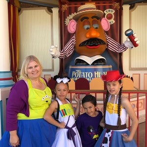 Puede incluir: Una familia vestida como personajes de la pel&iacute;cula Toy Story posa para una foto con el Sr. Cara de Papa en un parque de atracciones. La madre lleva un su&eacute;ter morado y un delantal amarillo con un dise&ntilde;o de extraterrestre. La hija de la izquierda lleva un vestido blanco con un dise&ntilde;o colorido y un lazo blanco en el pelo. La hija de la derecha lleva un sombrero de vaquero rojo, una camisa blanca, un cintur&oacute;n marr&oacute;n y una falda vaquera azul con estampado de vaca. El hijo lleva una camiseta morada con un dise&ntilde;o de Mickey Mouse. El personaje del Sr. Cara de Papa sostiene un micr&oacute;fono y lleva una camisa a rayas rojas y blancas y un sombrero de paja.