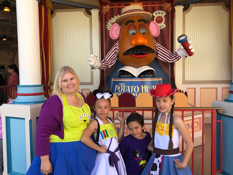 Puede incluir: Una familia vestida como personajes de la pel&iacute;cula Toy Story posa para una foto con el Sr. Cara de Papa en un parque de atracciones. La madre lleva un su&eacute;ter morado y un delantal amarillo con un dise&ntilde;o de extraterrestre. La hija de la izquierda lleva un vestido blanco con un dise&ntilde;o colorido y un lazo blanco en el pelo. La hija de la derecha lleva un sombrero de vaquero rojo, una camisa blanca, un cintur&oacute;n marr&oacute;n y una falda vaquera azul con estampado de vaca. El hijo lleva una camiseta morada con un dise&ntilde;o de Mickey Mouse. El personaje del Sr. Cara de Papa sostiene un micr&oacute;fono y lleva una camisa a rayas rojas y blancas y un sombrero de paja.