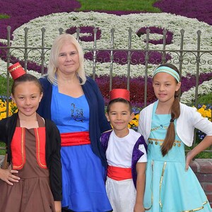 May include: A group of four people dressed in costumes from the movie Aladdin. The woman is wearing a blue dress with a red sash, the girl on the left is wearing a brown dress with a red vest, the boy in the middle is wearing a white shirt with a purple vest and red sash, and the girl on the right is wearing a turquoise dress with a white cardigan.