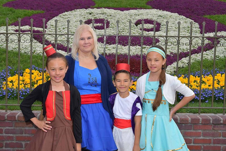 May include: A group of four people dressed in costumes from the movie Aladdin. The woman is wearing a blue dress with a red sash, the girl on the left is wearing a brown dress with a red vest, the boy in the middle is wearing a white shirt with a purple vest and red sash, and the girl on the right is wearing a turquoise dress with a white cardigan.