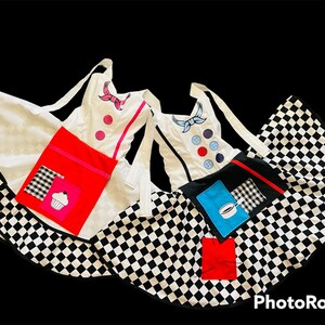 May include: Two black and white checkered aprons with white fabric tops. One apron has a red pocket with a pink cupcake design and the other has a blue pocket with a coffee cup design. Both aprons have a white fabric top with colorful embroidered details.