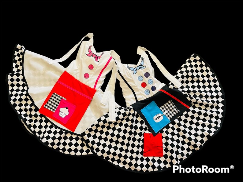 May include: Two black and white checkered aprons with white fabric tops. One apron has a red pocket with a pink cupcake design and the other has a blue pocket with a coffee cup design. Both aprons have a white fabric top with colorful embroidered details.