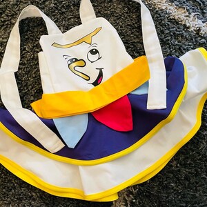 May include: A white apron with a yellow trim and a blue and white skirt. The apron features a cartoon face with a yellow smile and a blue nose.