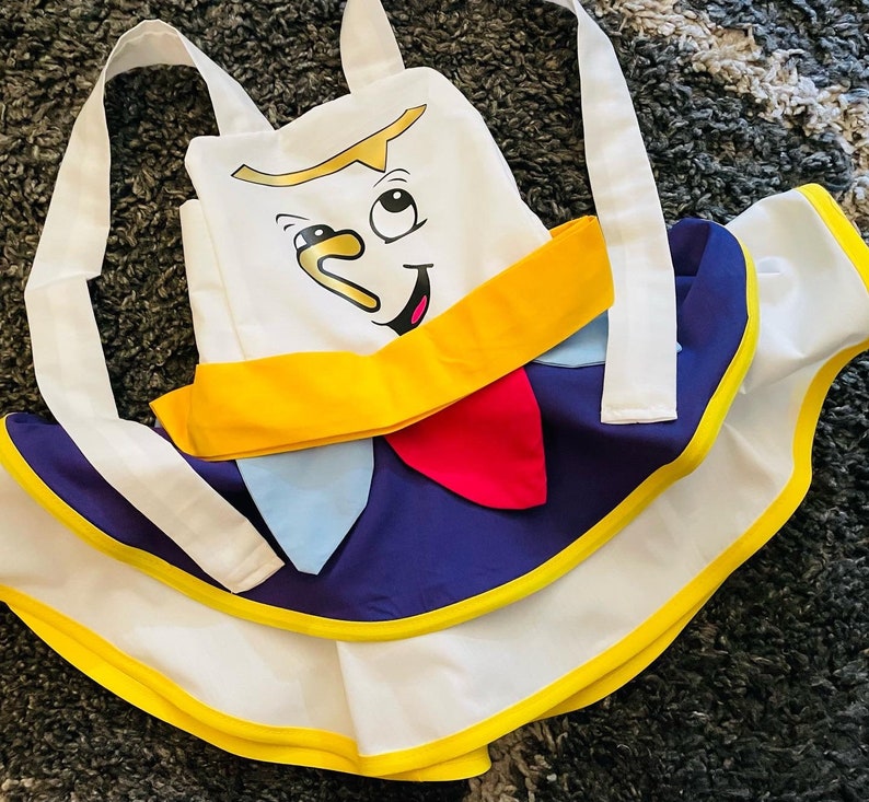May include: A white apron with a yellow trim and a blue and white skirt. The apron features a cartoon face with a yellow smile and a blue nose.