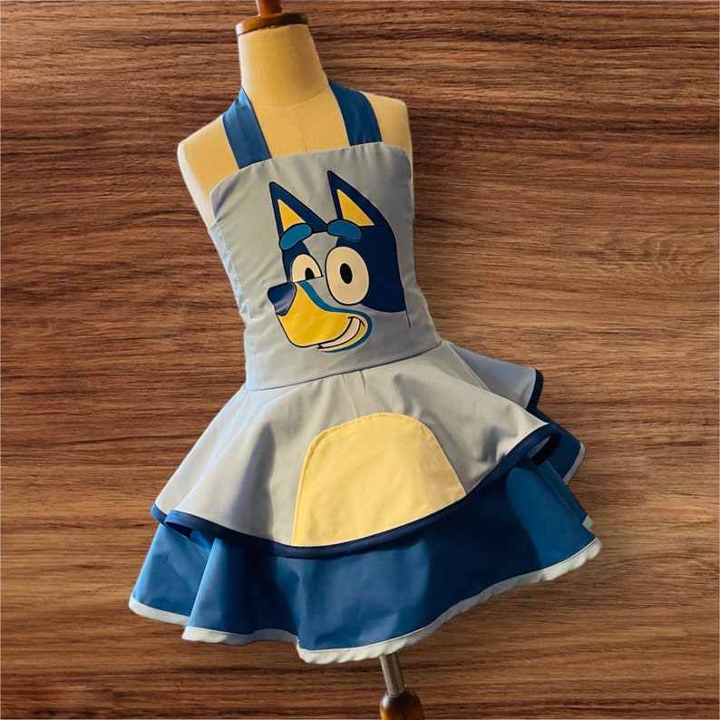 Op de afbeelding: Een blauwe en gele kinder schort met een cartoon hond ontwerp. Het schort heeft een geruite rok en een gele zak.