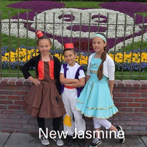May include: Three children dressed in costumes from the movie Aladdin. The girl on the left is wearing a brown and red costume with a red fez. The boy in the middle is wearing a white and purple costume with a red fez. The girl on the right is wearing a light blue dress with a gold belt and a white headband. The children are standing in front of a flower bed shaped like a Mickey Mouse head.