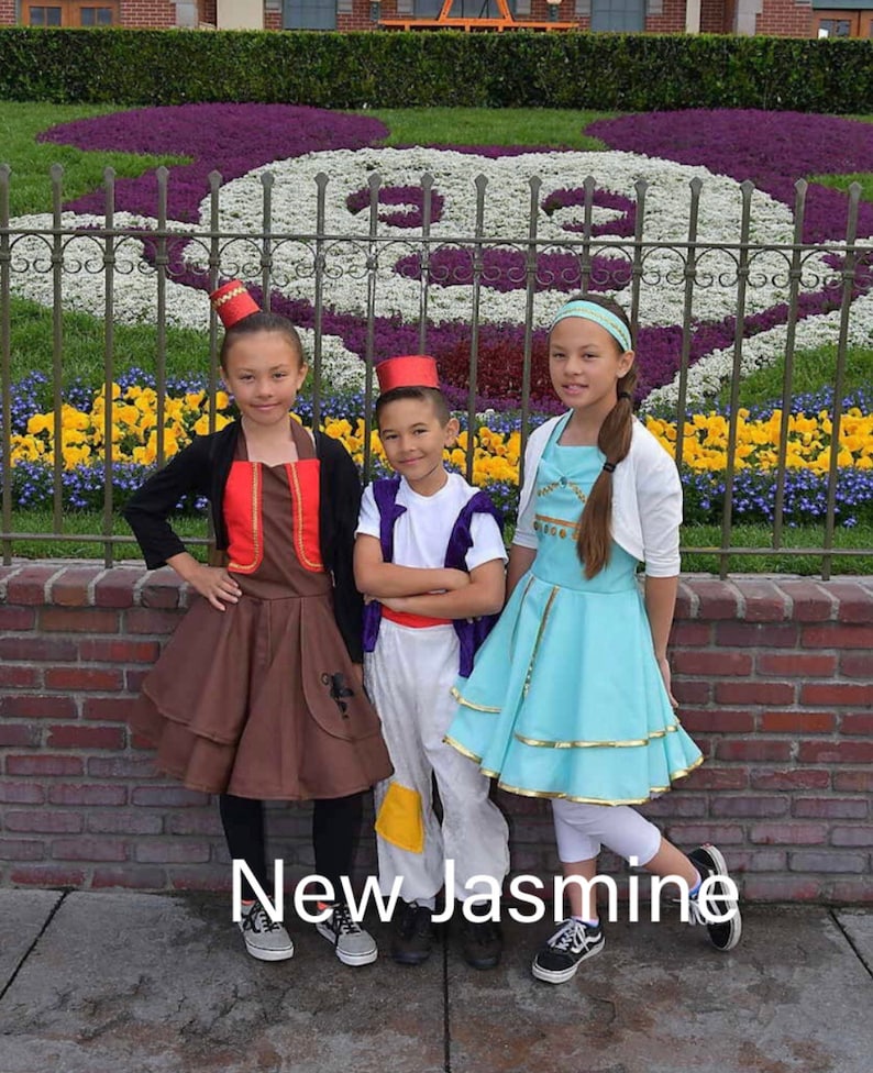May include: Three children dressed in costumes from the movie Aladdin. The girl on the left is wearing a brown and red costume with a red fez. The boy in the middle is wearing a white and purple costume with a red fez. The girl on the right is wearing a light blue dress with a gold belt and a white headband. The children are standing in front of a flower bed shaped like a Mickey Mouse head.