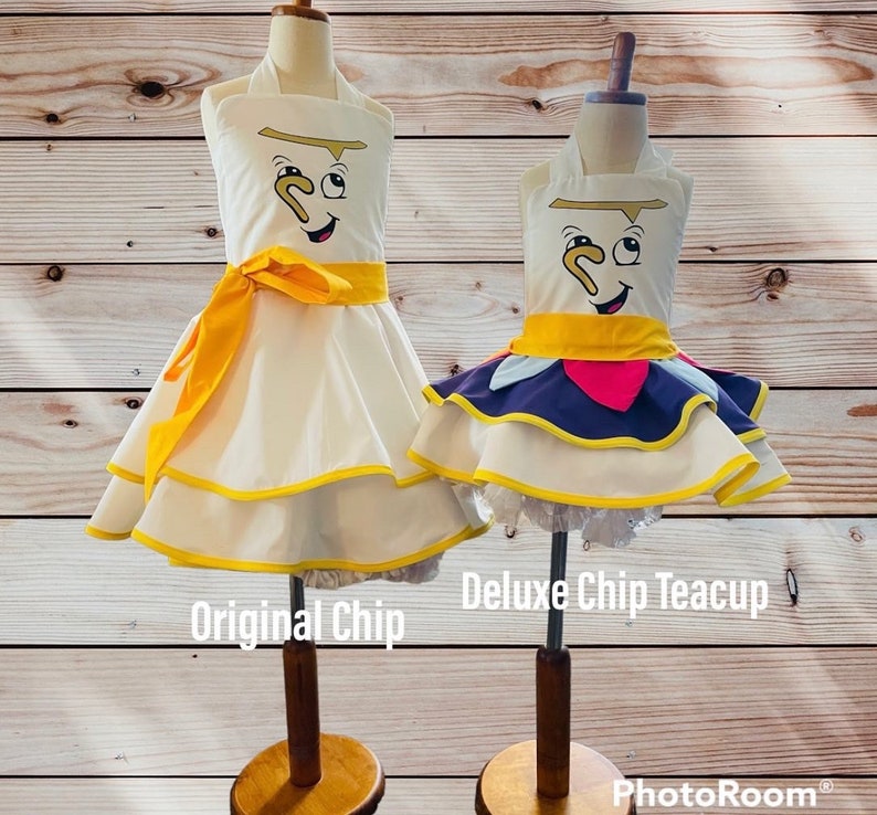 May include: Two Chip teacup costumes, one with a simple white skirt and the other with a purple, pink, and yellow layered skirt. Both costumes feature a yellow sash and a yellow trim around the skirt. The text "Original Chip" and "Deluxe Chip Teacup" is visible below the costumes.