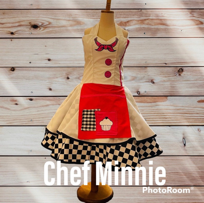 May include: A white and red chef apron with a black and white checkered skirt. The apron has a pink bow, two buttons, and a cupcake design. The text "Chef Minnie" is written below the apron.
