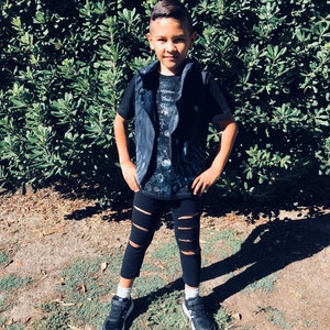 May include: A young boy wearing a black vest over a black and white graphic t-shirt, black ripped leggings, and black sneakers. He is standing in front of a green bush.