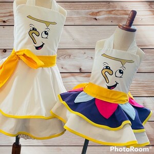 May include: Two white dresses with yellow sashes and a cartoon face of a teacup on the front. The dress on the left has a simple skirt, while the dress on the right has a layered skirt with purple, pink, and blue fabric.