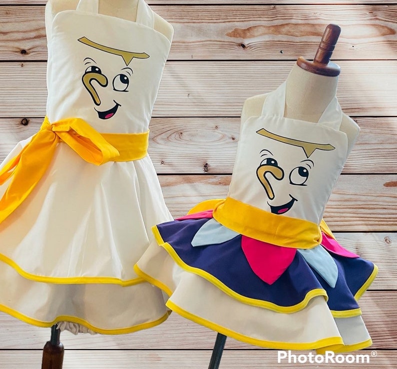May include: Two white dresses with yellow sashes and a cartoon face of a teacup on the front. The dress on the left has a simple skirt, while the dress on the right has a layered skirt with purple, pink, and blue fabric.