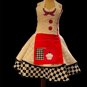 May include: A white and red dress with a black and white checkered skirt. The dress has a pink bow and two buttons on the bodice. The skirt has a red apron with a black and white checkered pocket and a pink cupcake graphic.
