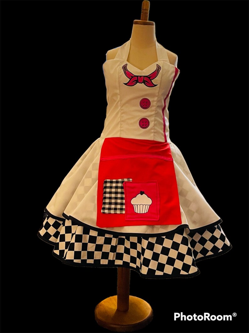 May include: A white and red dress with a black and white checkered skirt. The dress has a pink bow and two buttons on the bodice. The skirt has a red apron with a black and white checkered pocket and a pink cupcake graphic.