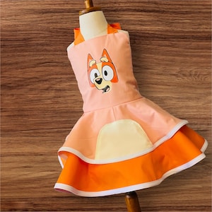 Op de afbeelding: Een roze en oranje kinder schort met een cartoon hondenkop op de voorkant. Het schort heeft een geruite rok en een witte rand.