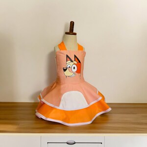 Op de afbeelding: Een roze en oranje jurk met een cartoonhond op het lijfje. De jurk heeft een witte rand en een gelaagde rok.