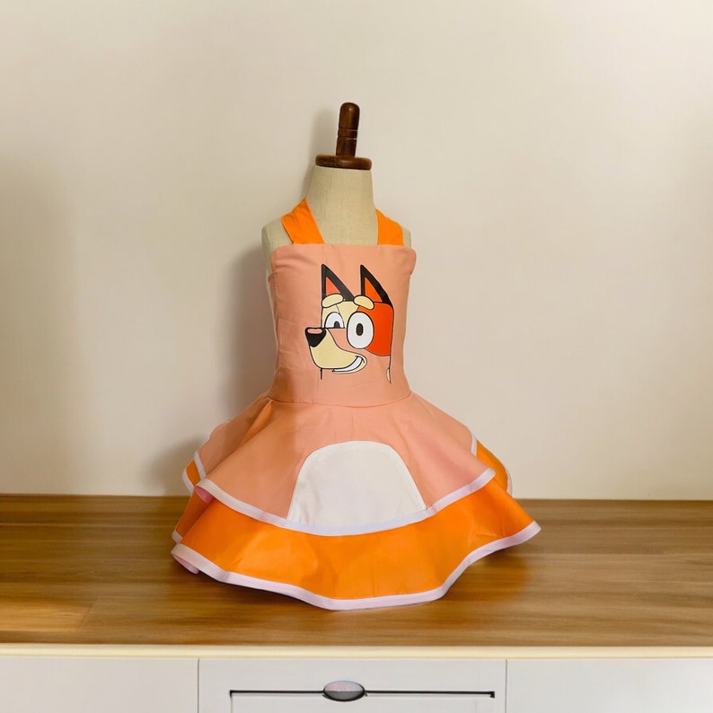 Op de afbeelding: Een roze en oranje jurk met een cartoonhond op het lijfje. De jurk heeft een witte rand en een gelaagde rok.