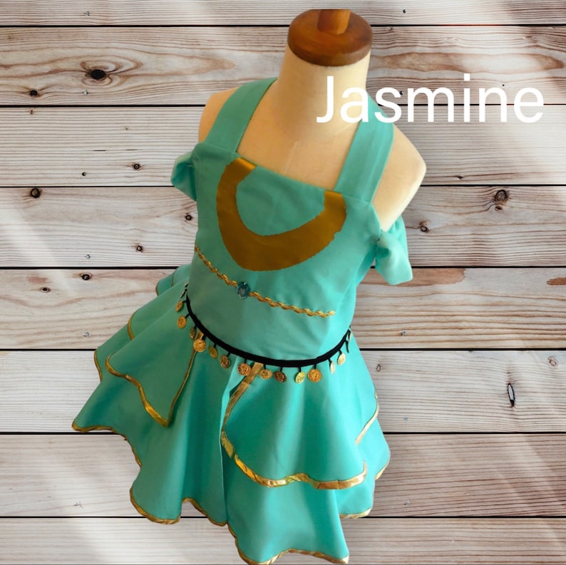 May include: A turquoise dress with gold trim and black and gold coin accents. The dress has a scoop neckline and a fitted bodice. The skirt is layered and has a ruffled hem. The dress is labeled "Jasmine".