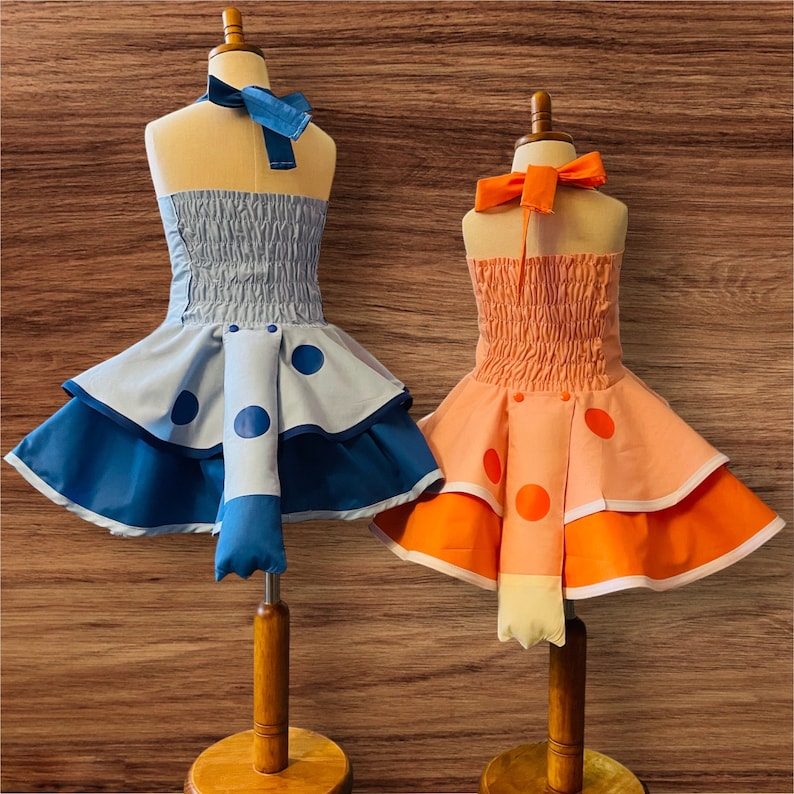 Op de afbeelding: Twee handgemaakte poppenjurken met staarten. De jurk links is blauw met witte stippen en een blauwe staart. De jurk rechts is perzikkleurig met oranje stippen en een perzikkleurige staart.