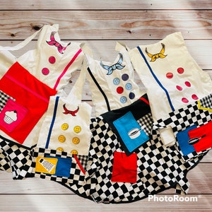 May include: Five white aprons with black and white checkered fabric and colorful patches. The patches include a cupcake, a grater, a cooking pot, a frying pan, and a button.