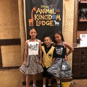 May include: Three children wearing animal print clothing pose in front of a sign that reads "Disney's Animal Kingdom Lodge". The sign features a giraffe and zebra illustration. The children are wearing black and white zebra print, brown and white giraffe print, and a yellow vest with a black animal print.
