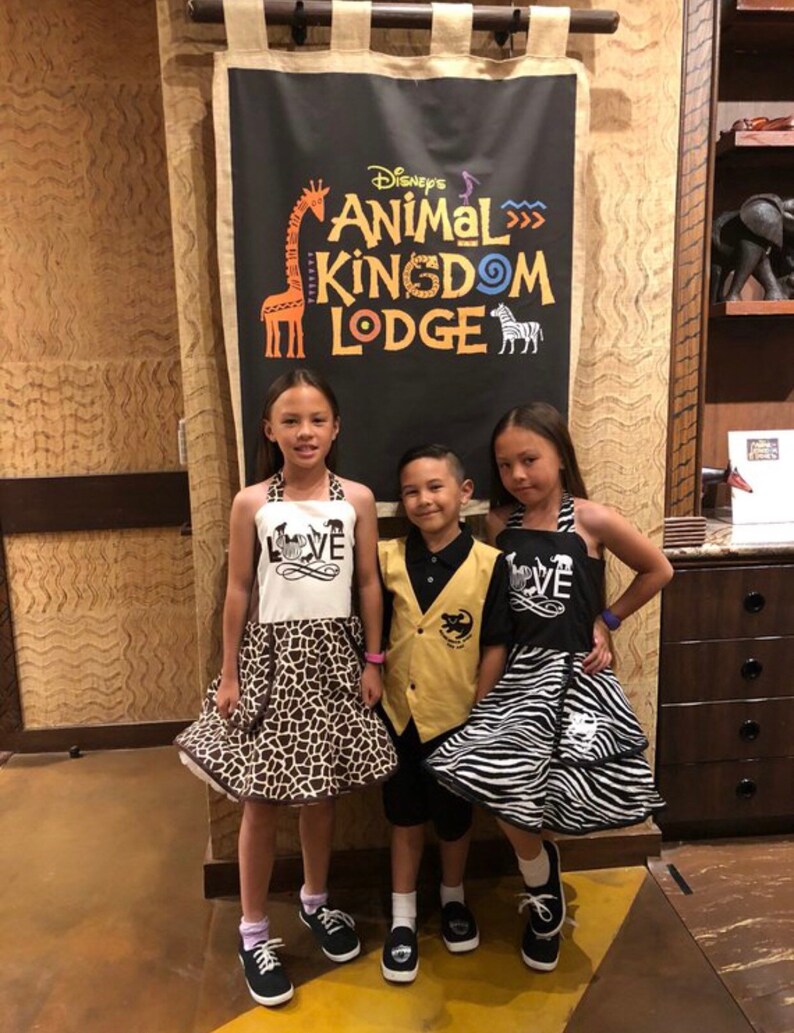 May include: Three children wearing animal print clothing pose in front of a sign that reads "Disney's Animal Kingdom Lodge". The sign features a giraffe and zebra illustration. The children are wearing black and white zebra print, brown and white giraffe print, and a yellow vest with a black animal print.