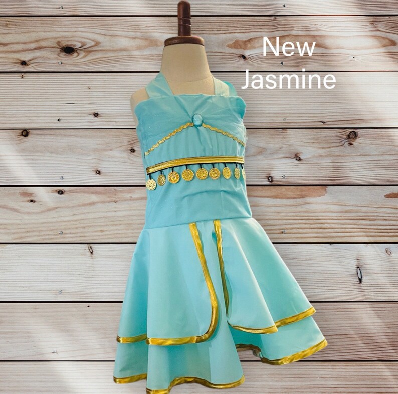 May include: A light blue dress with gold trim and coin accents. The dress has a halter neckline and a layered skirt. The text "New Jasmine" is written in white on a wooden background.