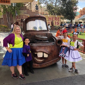 Puede incluir: Un grupo de personas disfrazadas de personajes de la pel&iacute;cula Cars de Disney Pixar, de pie frente a un gran cami&oacute;n de remolque de dibujos animados, marr&oacute;n, oxidado y de gran tama&ntilde;o. El cami&oacute;n de remolque tiene una gran sonrisa de dientes y un par de ojos grandes y azules. Las personas sonr&iacute;en y miran a la c&aacute;mara. El cami&oacute;n de remolque est&aacute; frente a un edificio con las palabras "Here It Is" en &eacute;l.