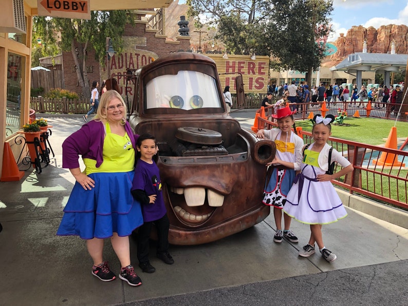 Puede incluir: Un grupo de personas disfrazadas de personajes de la pel&iacute;cula Cars de Disney Pixar, de pie frente a un gran cami&oacute;n de remolque de dibujos animados, marr&oacute;n, oxidado y de gran tama&ntilde;o. El cami&oacute;n de remolque tiene una gran sonrisa de dientes y un par de ojos grandes y azules. Las personas sonr&iacute;en y miran a la c&aacute;mara. El cami&oacute;n de remolque est&aacute; frente a un edificio con las palabras "Here It Is" en &eacute;l.