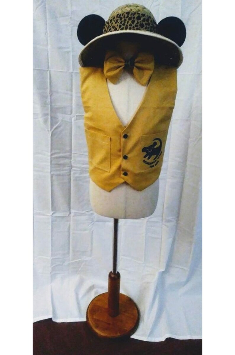 May include: A yellow vest with a pocket on each side and a black button closure. The vest has an embroidered image of a lion with the text "Remember You Are Brave" on the front. The vest is on a white mannequin with a brown wooden base. The mannequin is wearing a brown and black leopard print hat with black Mickey Mouse ears and a yellow bow tie.