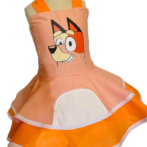 Op de afbeelding: Een roze en oranje jurk met een cartoon hondenkop op de voorkant. De jurk heeft een witte zak en een geruite rok.