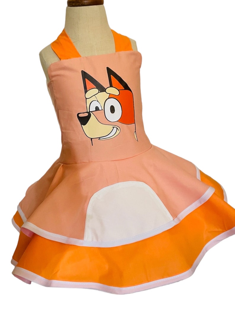 Op de afbeelding: Een roze en oranje jurk met een cartoon hondenkop op de voorkant. De jurk heeft een witte zak en een geruite rok.