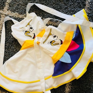 May include: Two white aprons with yellow trim and cartoon faces. One apron has a blue and yellow design with a cartoon face on the front. The other apron has a yellow trim and a cartoon face on the front.