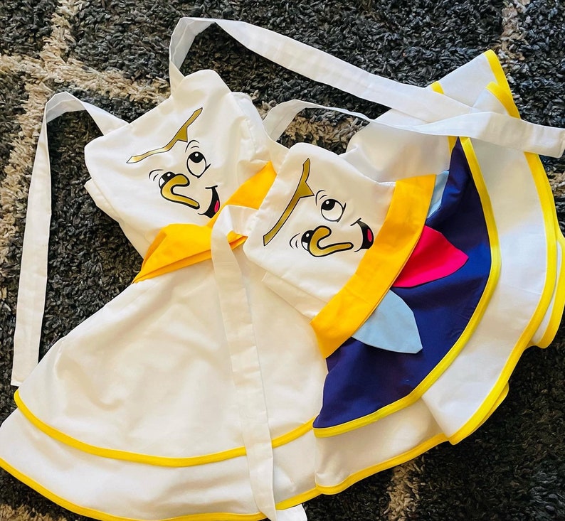 May include: Two white aprons with yellow trim and cartoon faces. One apron has a blue and yellow design with a cartoon face on the front. The other apron has a yellow trim and a cartoon face on the front.