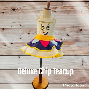 May include: A white teacup dress with a yellow, purple, and red ruffle skirt. The dress features a cartoon face of Chip, the teacup from Beauty and the Beast, with the text "Deluxe Chip Teacup".