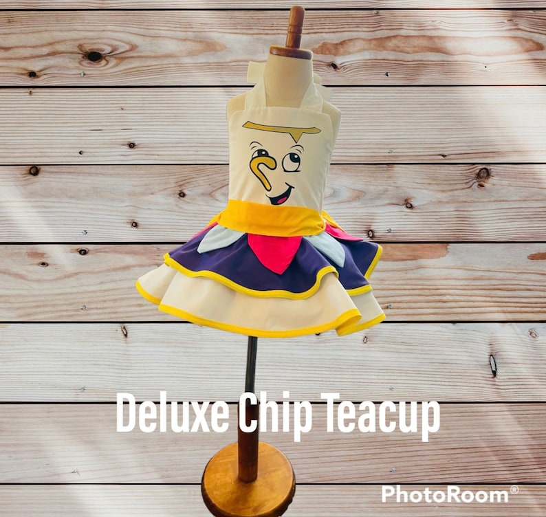 May include: A white teacup dress with a yellow, purple, and red ruffle skirt. The dress features a cartoon face of Chip, the teacup from Beauty and the Beast, with the text "Deluxe Chip Teacup".