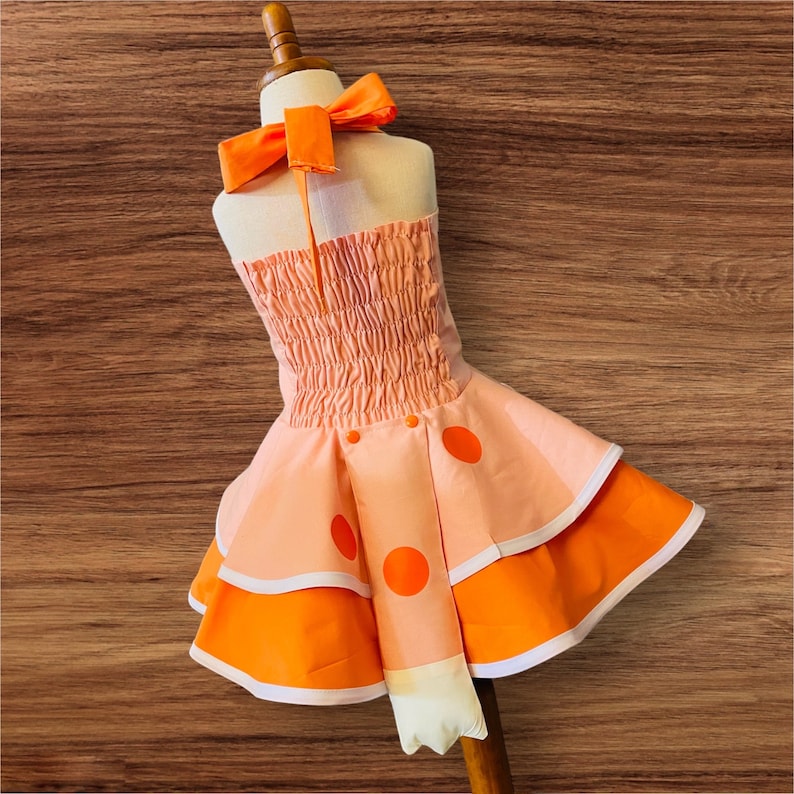 Op de afbeelding: Een roze en oranje jurk met een geruite rok en oranje stippen. De jurk heeft een haltertop met een strik.
