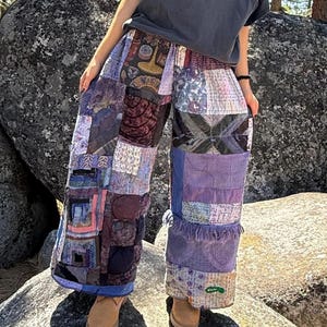 Peut inclure: Pantalon large à jambes larges présentant un motif patchwork composé de divers tissus dans des tons de violet, bleu et gris. Le pantalon présente des textures, des motifs et des bords effilochés variés, associé à un t-shirt gris foncé.