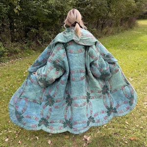 May include: A long, loose-fitting coat constructed from a vintage-style quilt featuring a double wedding ring pattern in shades of sage green, teal, and muted floral prints. The garment has wide sleeves, a large collar, and a flared, flowing hemline.