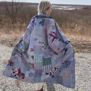 Może przedstawiać: Płaszcz patchwork w odcieniach niebieskiego, lawendy i różu. Płaszcz ma różne wzory i faktury tkanin, z widocznymi szwami. Płaszcz jest noszony na zewnątrz, z naturalnym krajobrazem w tle.