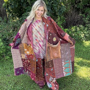 May include: A long, open-front coat made from patchwork fabric featuring various floral and geometric patterns in shades of burgundy, brown, orange, and teal. The garment includes two large front pockets and is worn over a tie-dye shirt and matching pants.