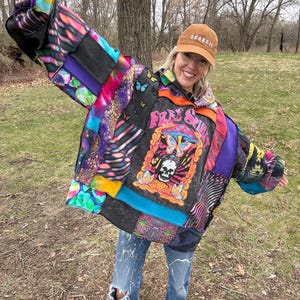 Op de afbeelding: Een kleurrijke patchworkjas met diverse patronen, waaronder tie-dye en bloemenprints. De jas heeft een afbeelding met een schedel, vlinders en de woorden "Free Spirit". De persoon draagt een bruine pet en een blauwe jeans.