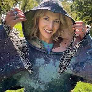 May include: A hooded poncho in shades of blue, green, and purple, with a starry pattern. The poncho has fringed edges and a lined hood. The person in the image is wearing the poncho and smiling.