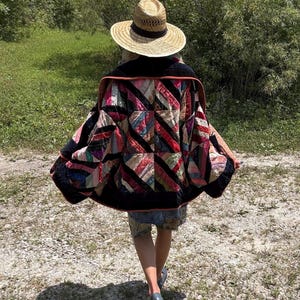 May include: A straw hat with a black band and a quilted jacket featuring a patchwork design of black, pink, red, and multi-colored fabric strips arranged in a geometric pattern with a dark collar and orange trim, worn over a blue shirt and shorts.