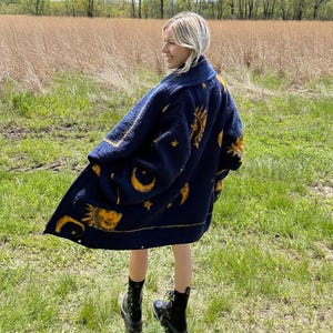 May include: A navy blue coat with a celestial design featuring suns, moons, and stars in yellow. The coat is worn outdoors in a grassy field, and the person is wearing black boots.