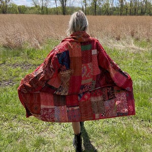May include: A patchwork red coat with a variety of patterns and textures. The coat is made of different fabric squares sewn together. The coat is being worn outdoors in a grassy field with trees in the background.