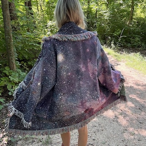 May include: A long-sleeved open-front cardigan featuring a woven galaxy print with deep shades of purple, blue, and black, accented by white star-like speckles. The garment is finished with a textured fringe trim along the collar and hemline.