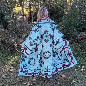 May include: A long, loose-fitting coat made from a light blue quilted fabric featuring a repeating pattern of red floral wreaths, dark green leaves, and dark red ribbon bows. The garment has wide sleeves and a wide collar with a dark green trim.