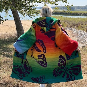 May include: A long-sleeved hooded cardigan featuring a vibrant rainbow gradient background with large, dark purple butterfly silhouettes printed across the fabric. The garment has a loose, open-front design with wide sleeves and a hood.