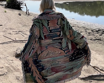 May include: A long, open-front woven tapestry coat featuring a multi-colored landscape scene with a large sun, dark sky, and green foliage. The garment has a wide collar, long sleeves, and frayed edges along the hem and cuffs, creating a textured look.